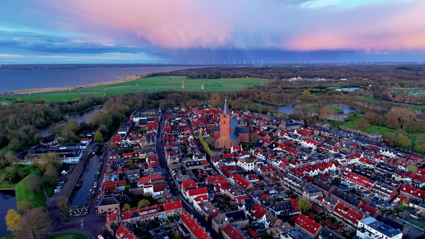 A magnificent wide-angle aerial photograph capturing a sweeping panoramic sunset view over a historic medieval fortress town. The lower half of the frame highlights a dense, beautifully organized grid of traditional European houses with distinct red-tiled roofs and narrow streets, centering around a massive brick church cathedral with a tall, sharp spire. In the background, the flat landscape extends to defensive water moats, green polder fields, and a massive blue river delta or coastal bay. The distant horizon features a long line of modern industrial wind turbines spinning under an extraordinarily dramatic sky, where massive storm clouds are illuminated in vibrant shades of pink, orange, and deep purple by the setting sun. This high-resolution capture perfectly documents ancient urban architecture and modern green energy infrastructure.