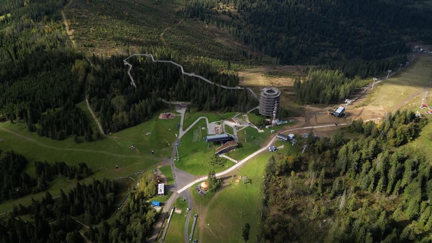 Aerial view of a winding elevated treetop walkway through dense forest, with open grassy slopes and a mountain park setting in summer light


