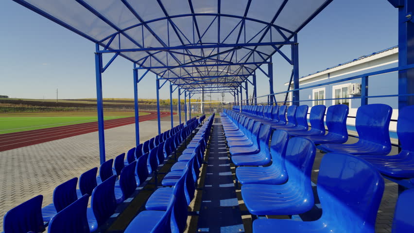 Active view reveals row of plastic seats overlooking football field and running track. Sunlight illuminates empty stand hiding under canopy