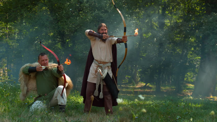 Two Dacian reenactors shoot flaming arrows with bows in a sunlit woodland clearing.