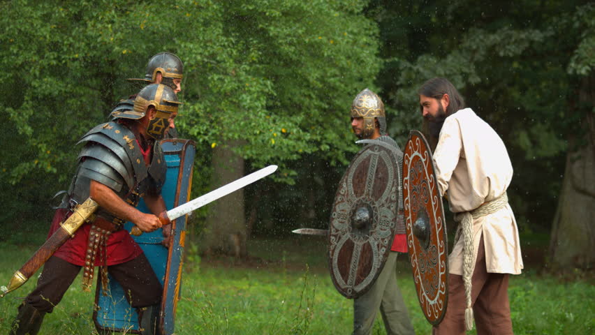Roman soldier confronts Dacian warriors with shields and swords during historical reenactment in a green woodland.