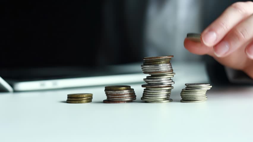 Person stacks coins on a table while using a smartphone in a modern office space during the day.