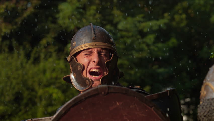 Close-up of Roman legionary in helmet yelling behind shield during rainy battle reenactment in forest.
