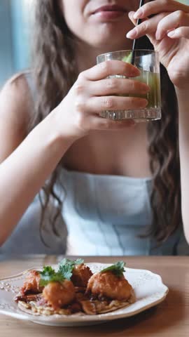 Young woman smiles while stirring pale drink in short glass at dining table with plated food in foreground, indoor close view with soft natural light, blurred background, warm wood surface, relaxed