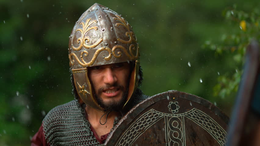 Close-up of a Dacian warrior in ornate helmet and chainmail raising a shield during a rainy battle reenactment.