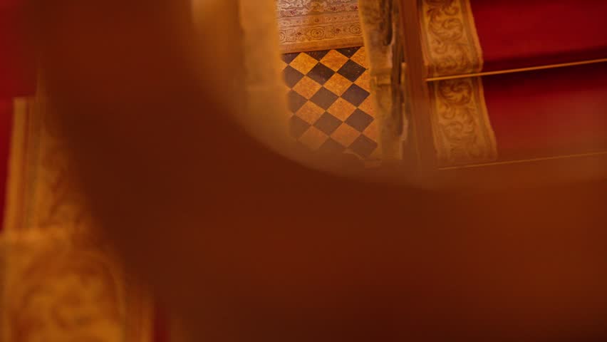 Warm, cinematic shot revealing a luxurious and historic hall with an antique wooden chair, a checkered floor, and rich red textiles. The view emerges from behind an ornate column or furniture