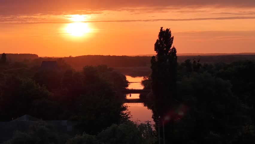 The drone hovers over weeping willows and wild riverside vegetation as the sun sinks toward the horizon, flooding the sky with amber and gold. A tall industrial chimney and low town rooftops break the skyline, a road bridge crosses the slow river, and birds glide through the warm light.