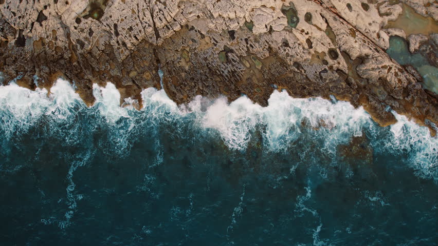 Aerial view of ocean waves crashing onto a rugged rocky shoreline in slow motion