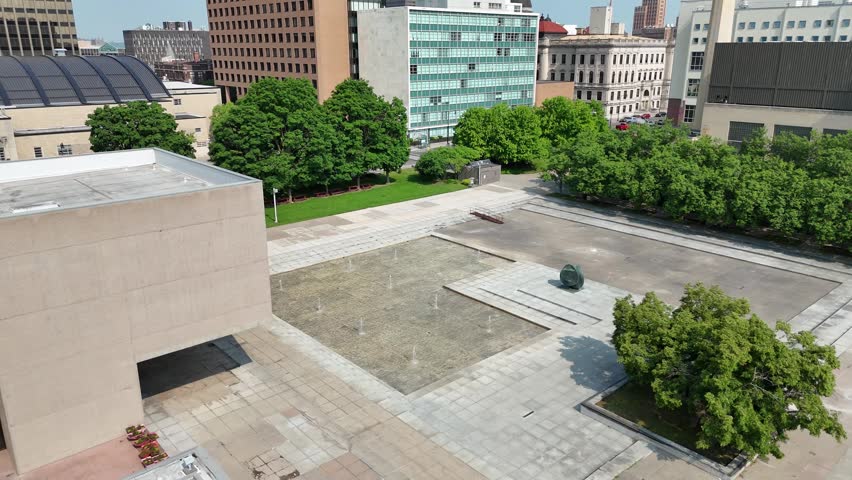 An open plaza and a large, shallow pool with numerous water jets dispensing water into the air sits adjacent to a building dedicated to being a museum on a beautiful sunny summer day.