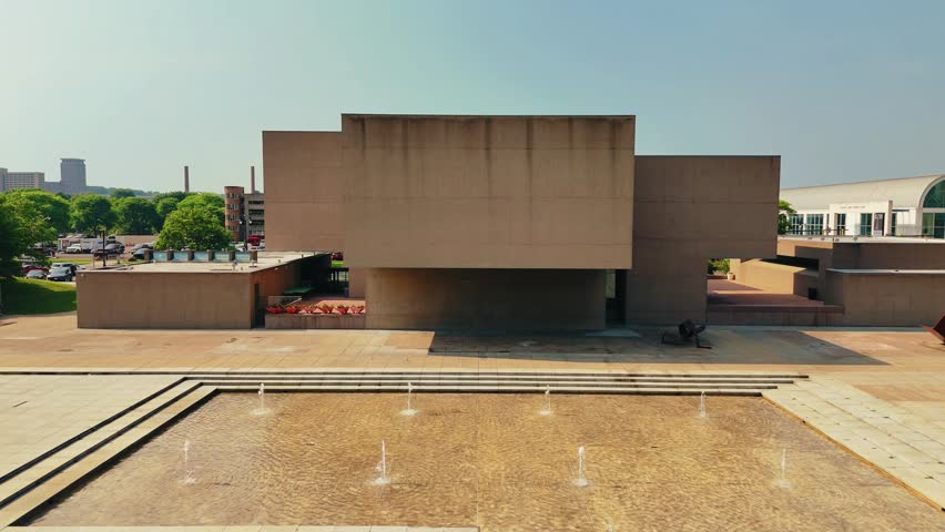 Water jets thrust water into the air from within a shallow pool that sits accompanying a museum within the middle of a city.