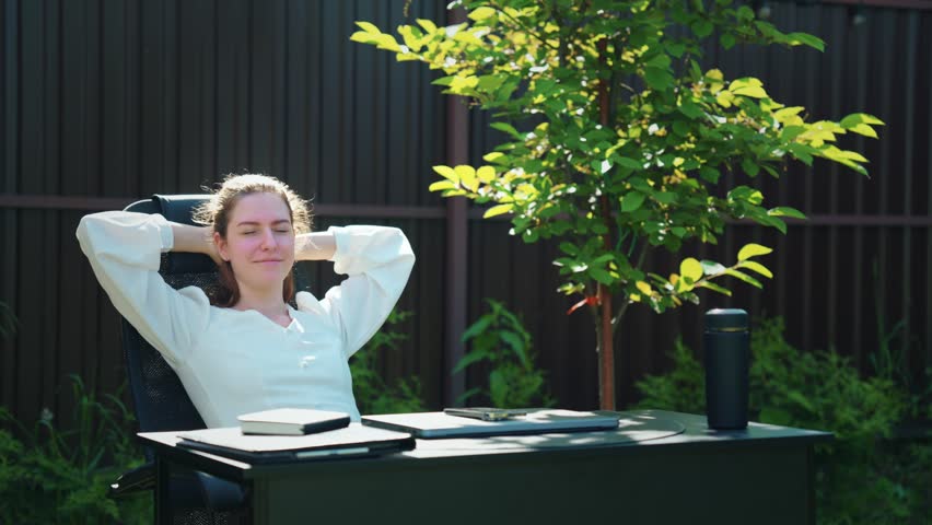Young businesswoman leaning back and relaxing at an outdoor workspace in a private garden, representing work break, remote work, healthy work-life balance and flexible digital lifestyle.