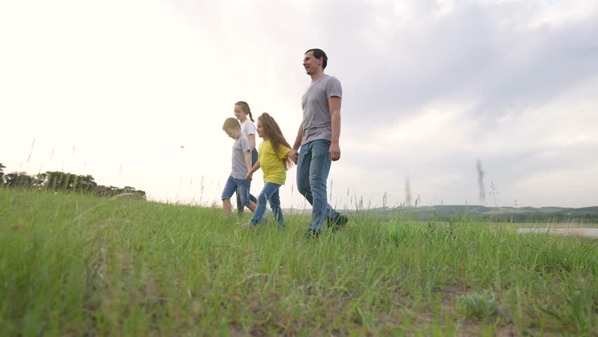 Family walks through green nature. Parent and child walk together. Everyone appears happy outside. People step across tall grass. Group feels calm by lake water. Family hikes over summer grass.