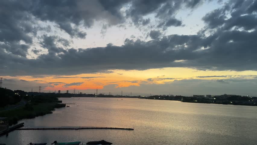 A dramatic cloudscape dominates the sky above a calm lake at dusk, with a warm orange and golden glow along the horizon beneath layers of dark blue and grey clouds. City lights begin to emerge along both shorelines, reflecting softly on the still water surface. Moored boats and a wooden dock are visible in the lower foreground, with power line towers silhouetted against the glowing sky in the distance.