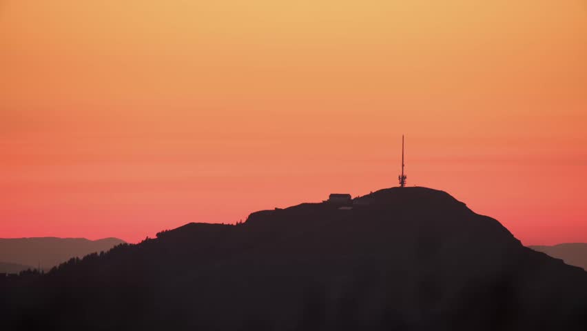 The sun sets behind the mountain, casting a warm glow. A communication tower stands tall against the colorful sky. Nature shows its beauty at dusk.