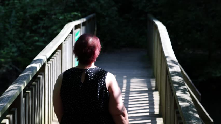Overweight woman with red hair walking across a wooden bridge in a forest and looking at the view
