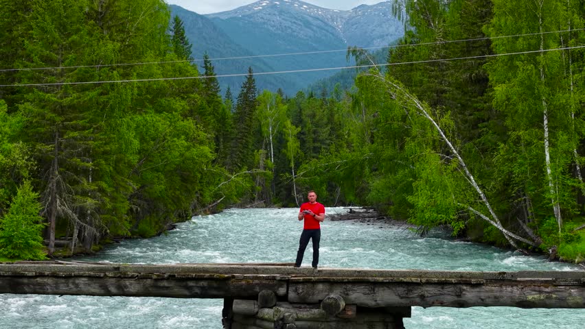 An athletic male tourist standing on a wooden bridge over a fast-flowing mountain river, holding a remote control and showing a peace sign.