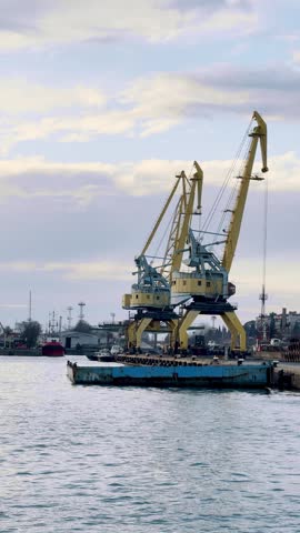Cranes working at a port during the day. Cranes lift containers at a busy port on a cloudy day with boats nearby and city buildings in the background.