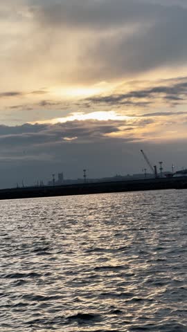 Cranes at sunset by water. Waves move gently as the sun sets behind clouds. Cranes stand tall at the harbor in the distance.