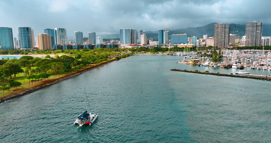 Flight over the waterscape surrounded by green park and multiple yachts at the shore. Skyline of Waikiki, Honolulu, Oahu, Hawaii, USA at backdrop.