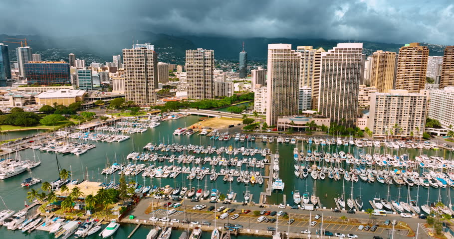 Flying to the yacht club with multiple luxurious vehicles at the berths. Approaching high-rise buildings of Waikiki in Honolulu, Hawaii, USA. Rainy clouds covered the cityscape at backdrop.