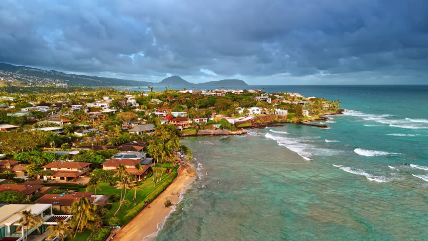 Beautiful villas in the stunning lush greenery on the picturesque shore of Hawaii, USA. Waves roll to the sandy beach. Heavy cloudscape covering the sky above the panorama. Aerial perspective.