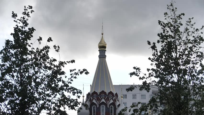 Golden spire of a Russian Orthodox church against a dramatic cloudy sky, framed by trees in an urban landscape.