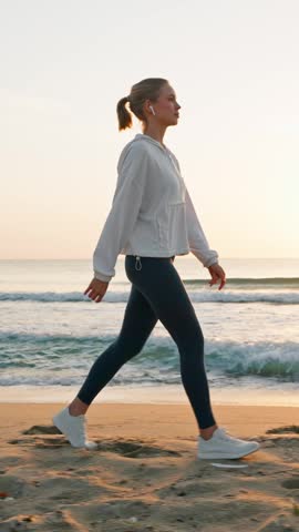 Young fit woman walking fast on sandy beach at sunrise vertical smartphone video orientation female athlete in wireless earphones exercising near sea waves morning workout healthy lifestyle.