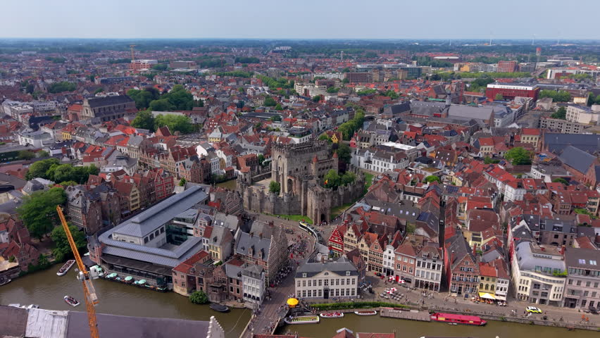 A magnificent wide-angle aerial photograph capturing the iconic Gravensteen castle (Castle of the Counts) situated in the historical center of Ghent. The frame highlights the sprawling medieval stone fortress with its defensive walls, towers, and courtyards, bordered by a scenic river canal where tour boats and pleasure craft are docked. In the foreground, an old fish market building with a grand arched roof sits right by the water, leading into a bustling public street filled with pedestrians. The fortress is enveloped by a dense, picturesque urban landscape of traditional Flemish townhouses and residential quarters featuring characteristic red and dark tiled roofs. In the background, the vast city grid extends to a flat summer horizon under a bright sky. This high-altitude overview perfectly documents medieval military architecture, heritage preservation, and regional tourism.