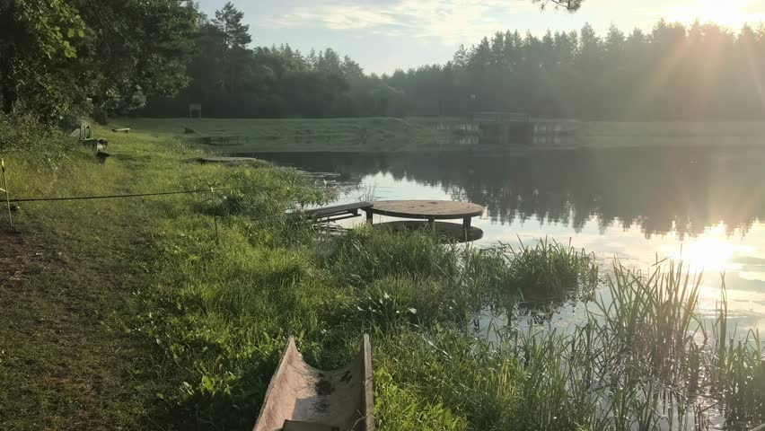 Two fishing poles are placed near the edge of a lake with green grass and plants around. The water is still and reflects the sky above while it is daytime.