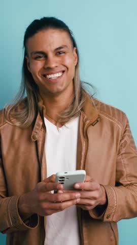Young happy man with long dyed hair using a smartphone and smiling, isolated on a light blue studio background. Positive male person chatting. Vertical
