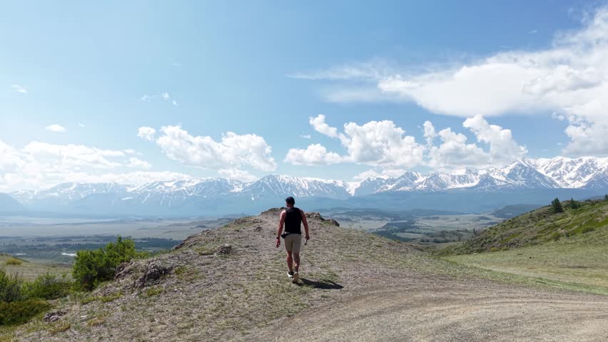 Young male traveler from behind walking forward along a rugged trail toward a majestic snow-covered mountain range under a cloudy sky