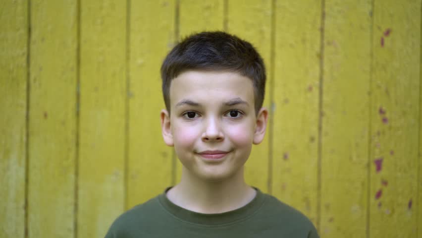 Cute young boy close-up portrait looking at camera with slight smile against yellow wooden wall. Green t-shirt, natural light, innocent expression.