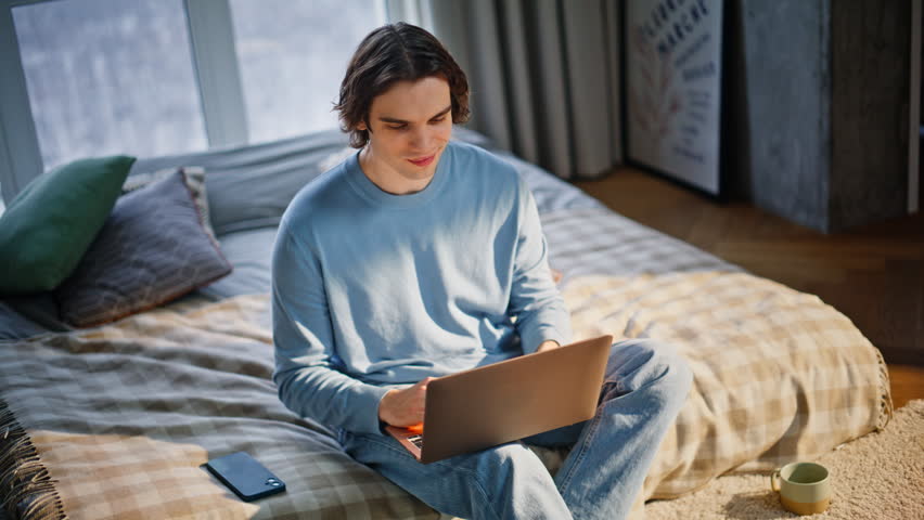 Smiling man video calling by laptop at comfortable apartment mattress closeup. Relaxed handsome guy have friendly online meeting home. Positive young male talking to computer webcam in sunny bedroom