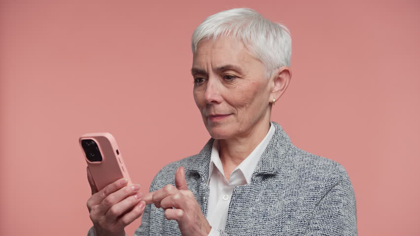 Surprised senior woman taps her smartphone while browsing online, reacting with curiosity, concern, and focus against a pink background
