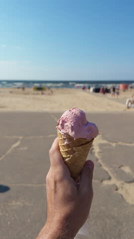 Hand holding pink ice cream cone on sunny beach promenade with sea and blurred summer visitors