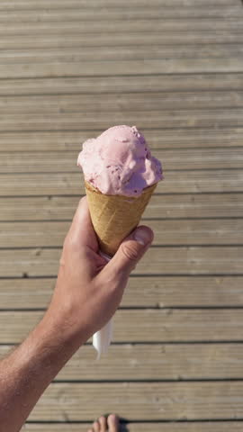 Hand holding strawberry ice cream cone on sunlit wooden boardwalk with barefoot step