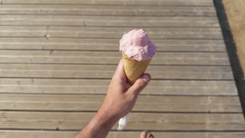 Hand holding pink strawberry ice cream cone over sunlit wooden boardwalk