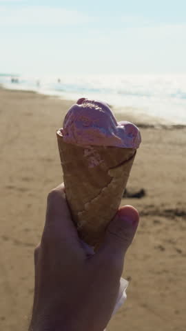 Hand holding melting pink ice cream cone on sunny beach with bright sea horizon