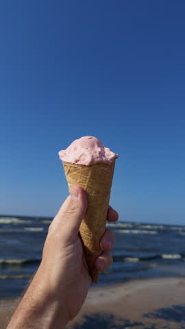 Hand holding strawberry ice cream cone against blue sky above seaside beach and ocean horizon