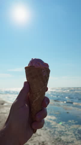 Hand holding ice cream cone with bright sun, blue sky and ocean shoreline on summer beach