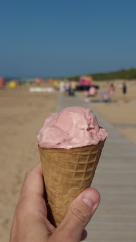 Hand holding pink ice cream cone on sunny beach with blurred seaside promenade and blue sky