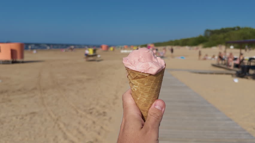 Hand holding strawberry ice cream cone on sunny beach boardwalk with blurred summer crowd