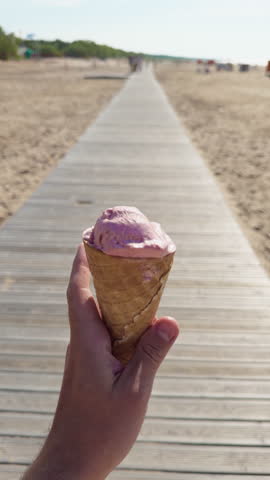 Hand holding pink ice cream cone on sunny beach boardwalk with blurred shoreline and open summer background