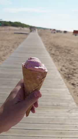 Hand holding pink ice cream cone on sunny beach boardwalk with blurred sandy shore and distant visitors