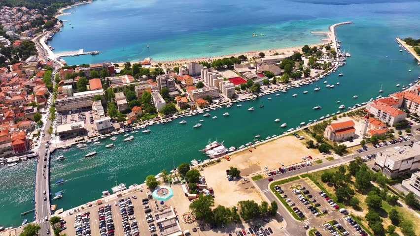 Omis shows a waterfront area with boats in the water. Nearby buildings and a beach are visible. A parking lot is located on the right side. People are present along the shore.