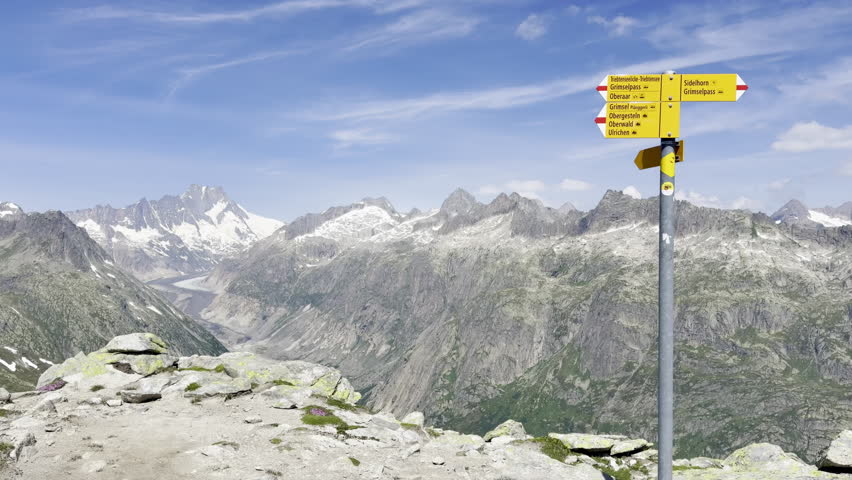 Yellow hiking signpost to Sidelhorn and Grimselpass above a high-alpine panorama of snow-capped Bernese Alps peaks and a glacial valley