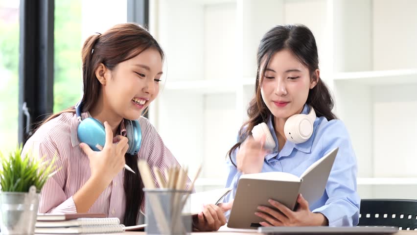 Two happy young Asian female students with headphones studying together, smiling and writing in a notebook at a desk. Education and lifestyle concept.