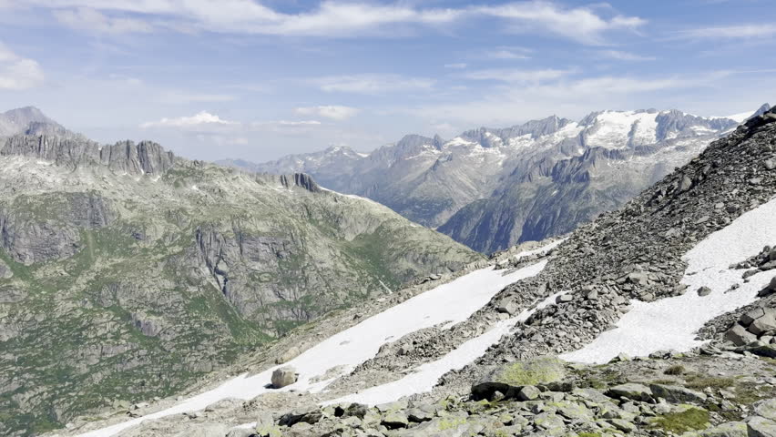 Hiking along a rocky ridge trail above the Grimsel Pass toward a summit cairn, overlooking a braided glacial river valley, alpine tarn and snow-capped Bernese Alps peaks, Switzerland