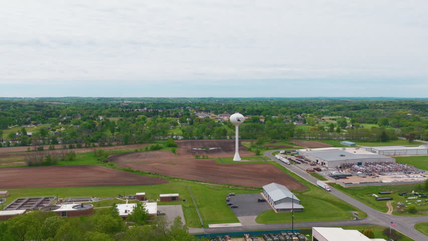 Drone view of the Berlin, Wisconsin water tower rising between freshly tilled farm fields, an industrial park with warehouses and semi trucks, and the tree-lined Fox River valley in spring.