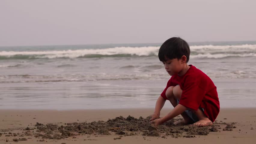 Child in red shirt sits barefoot while playing on the sand, Sunset bathes beach in soft, warm tones